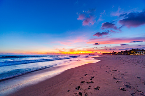 A serene beach at sunset. The orange and pink sky is reflected in the deep blue ocean with white waves crashing onto the shore. The light tan sand is scattered with footprints. In the background, there are a few buildings on the right side of the image. The photo is taken from a low angle, making the beach and ocean appear vast and expansive.
