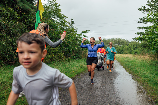 A group of children and their parents taking part in a fun run in a public park in Cramlington, North East England on a rainy day. The main focus is a woman crossing the line excitedly with her arms up after completing the run while looking at the volunteer who is standing at the finish waiting for them with her hand out, ready to hi-five the participants.\n\nVideos are also available for this scenario.