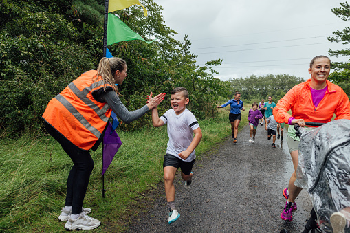 A group of children and their parents taking part in a fun run in a public park in Cramlington, North East England on a rainy day. The main focus is a boy crossing the finish line hi-fiving the volunteer who is standing at the finish waiting for them all to finish with her hand out.

Videos are also available for this scenario.