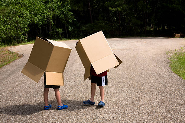 Two boys outside, road with large cardboard boxes over bodies. Two boxes playing "talking boxes" outdoors. Legs seen coming out of the large cardboard boxes. Street. Children.  two heads are better than one stock pictures, royalty-free photos & images