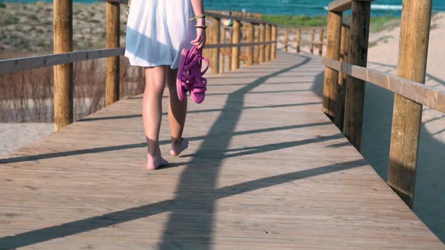 Close-up of a young woman walking towards the beach in a white dress holding sandals