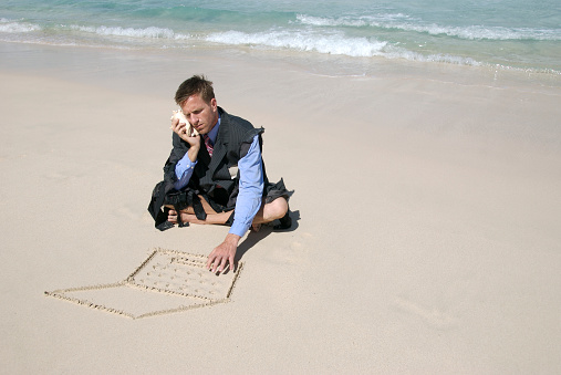 Castaway businessman types on his sand laptop and talks on his shell phone on a deserted beach