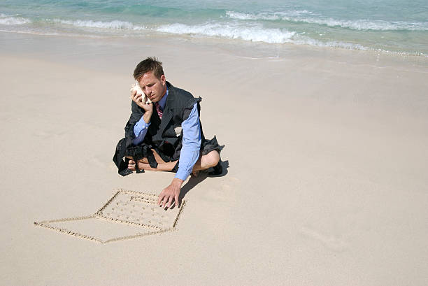 Castaway Businessman Multitasks Castaway businessman types on his sand laptop and talks on his shell phone on a deserted beach castaway stock pictures, royalty-free photos & images