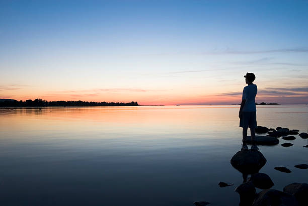 Young man standing on a lakeshore at sunset looking beyond stock photo