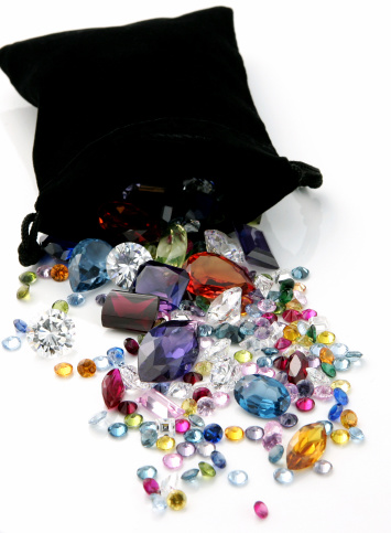 Colored gems and gemstones spilling out of black bag onto white.