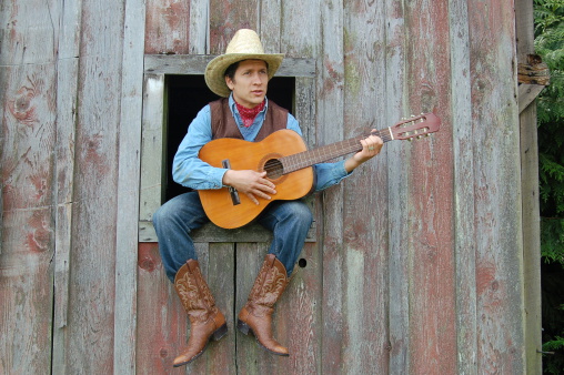 This cowboy plays his guitar while sitting in the window of his barn.