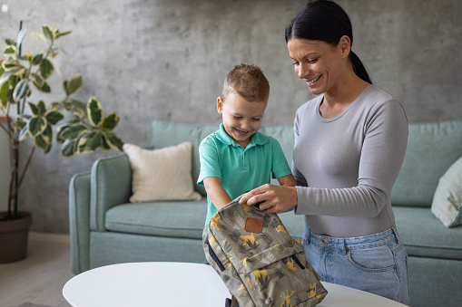 A mid adult Caucasian babysitter and a boy that she's taking care of packing a backpack for school in the living room of an apartment