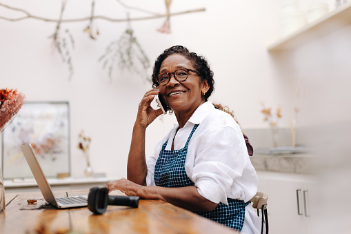 Happy flower store owner talks with her clients on a phone call, using her communication skills to build strong relationships with her customers. Senior craftswoman running a small business.