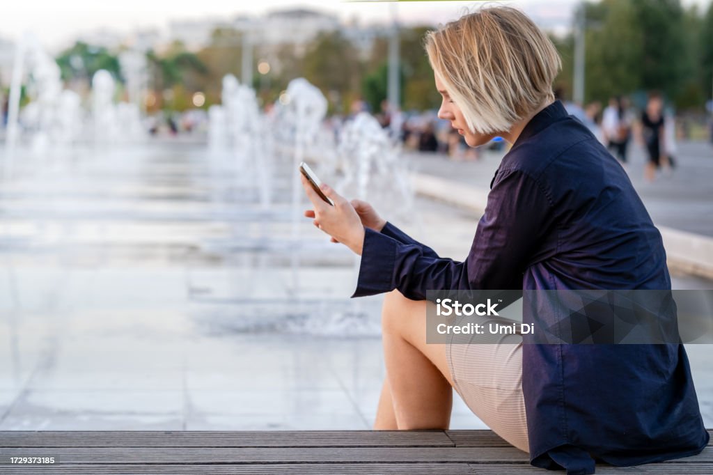 Walking in Thessaloniki. Teen girl sitting at the fountain and scrolling through the information in the phone on the embankment Teenage girl sitting at the fountain and scrolling through the information in the phone 14-15 Years Stock Photo