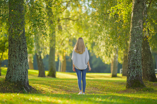 Young adult blonde woman slowly walking on green grass at city park in beautiful warm sunny autumn day. Spending time alone and enjoying freedom at nature. Peaceful atmosphere. Back view.