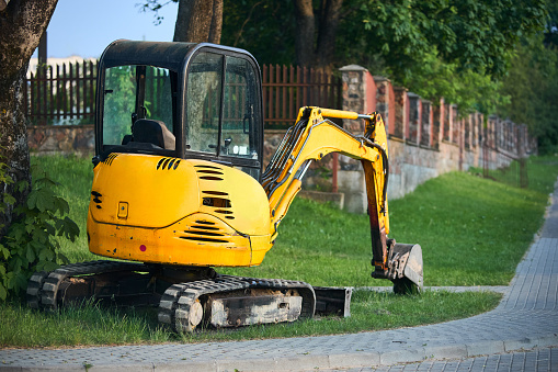 Tracked orange mini excavator digger near road with green trees and stone fence on background, with copyspace.