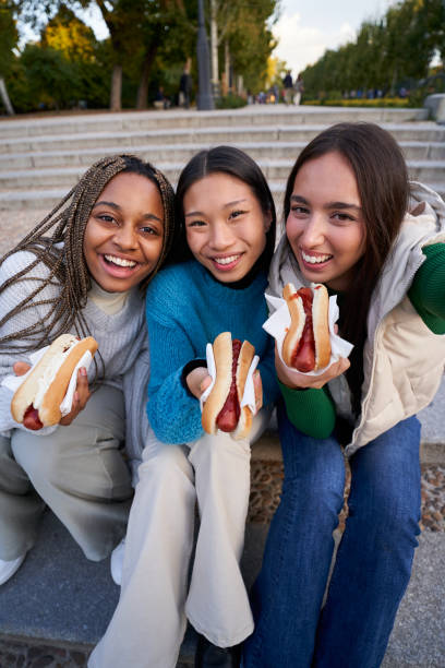 Three women student friends taking a selfie together while eating hotdogs outdoors. Group of three young female friends sitting on park stairs and eating hotdogs. Mixed race student ladies having fun at the city while eating fast food outdoors. Vertical selfie. selfie madrid photos stock pictures, royalty-free photos & images