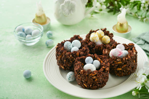 istock Easter chocolate nest cake with mini chocolate candy eggs with blossoming cherry or apple flowers on green background table. Creative recipe for Easter table with holiday decorations. Top view. 1729481119