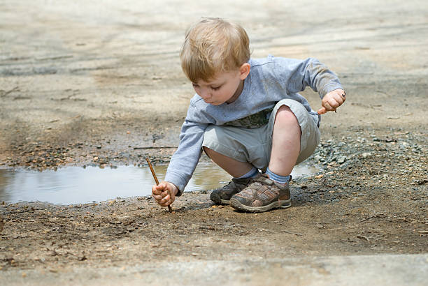 A small boy stoops near a mud puddle and plays quietly and intently with a stick.