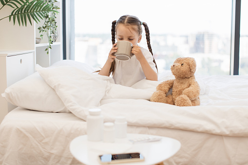Tired caucasian girl taking sip of hot tea to easing sore throat while warming herself in cozy bed. Cute child sitting next to teddy bear that supports her during cold indoors.
