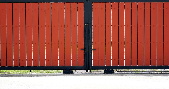 Front veiw of a red wooden slats Sliding gate with a black metal handle in the from of a fence.