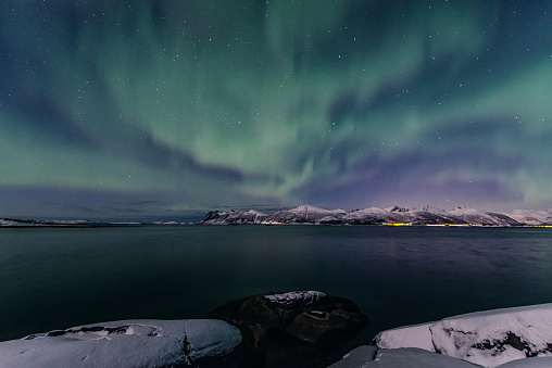 Northern Lights, polar light or Aurora Borealis in the night sky over Senja island in Northern Norway. Snow covered mountains in the background with water of the Norwegian Sea reflecting the lights in the foreground.