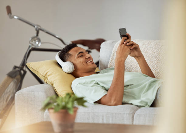 fones de ouvido, vídeo do telefone e homem em um sofá da sala de estar feliz e relaxar em casa. série, aplicativo engraçado e rindo com música e ouvindo em um salão da casa com web streaming móvel e sofá com sorriso - mobile phone home video camera video photograph - fotografias e filmes do acervo