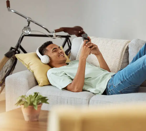 Photo of Headphones, phone video and relax man on a living room sofa happy at home. Series, funny app and laughing with music and listening in a house lounge with web mobile streaming and couch with smile