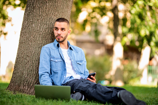 Young man using laptop for studying or working outdoor. Young male wearing casual clothes and sitting on grass in the public park and text messaging. Full length shot.