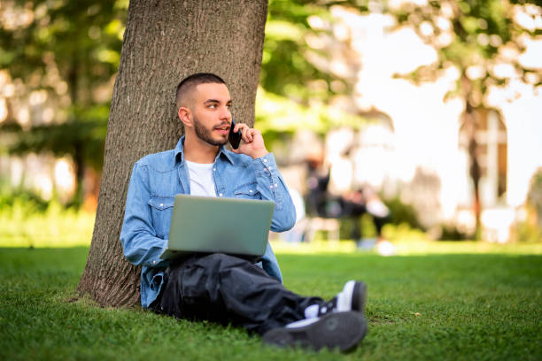 Full length shot of a young man using laptop and smartphone in the public park stock photo
