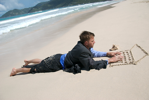 Castaway businessman mixes business with pleasure as he lays on a deserted beach working typing on his sand laptop