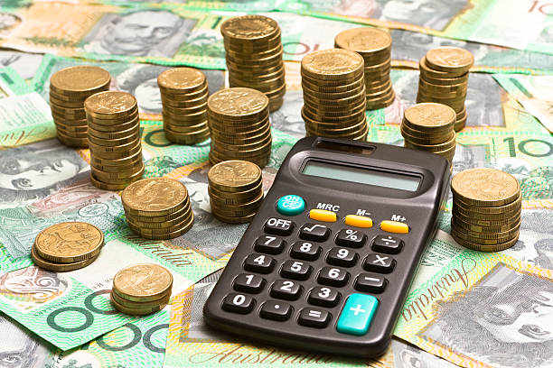 Adding up the profits "Calculator and coins on one hundred dollar notes. Doing the budget, adding up profits...." australian dollar stock pictures, royalty-free photos & images