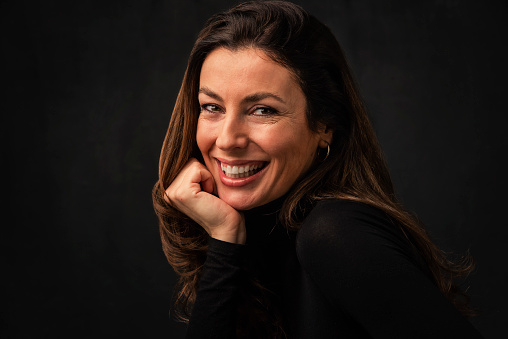 Headshot of an attractive middle aged woman with toothy smile wearing turtleneck sweater while sitting at isolated dark background. Copy space. Studio shot. Hand on forehead.