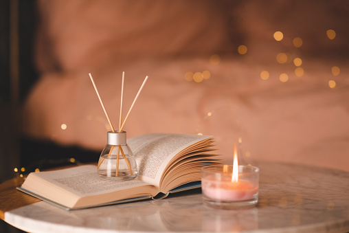 Liquid home fragrance in glass bottle with bamboo sticks and scented candle with open paper book on marble coffee table at bedroom over glow lights. Cozy home atmosphere. Aromatherapy.