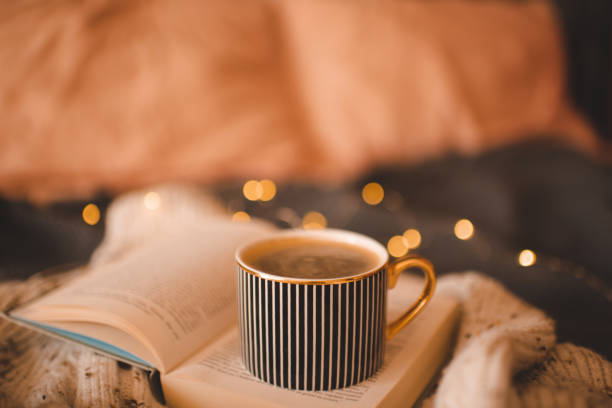 Cup of coffee on open paper book on knitted textile in bed over Christmas lights close up. Winter holiday season. Home hygge cozy atmosphere.