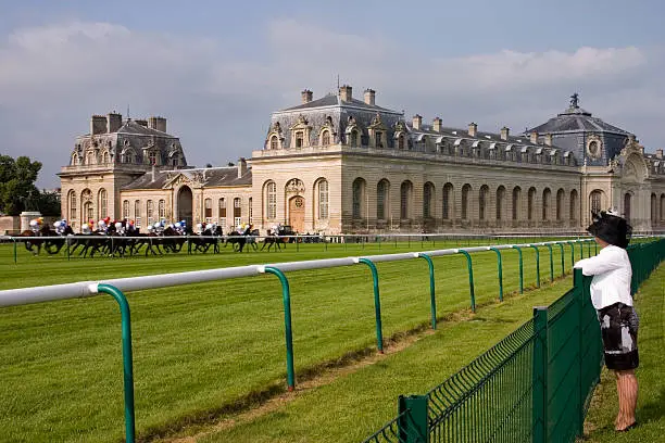 "On the public parkland at the stylish and fashionable horse racing centre of Chantilly, France, racing takes place in front of the Living Horse Museum, Le Musee Vivant du Cheval, which houses the worldaas most beautiful stables, and probably the largest at 186mm long. They were built in 1719, could house up to 240 horses and belonged to the adjacent, Chantilly Chateau which was in the ownership of the Princes de CondA. PLEASE NOTE: The pattern of the fabric on the ladies dress has been bastardised."