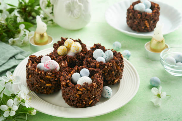 istock Easter chocolate nest cake with mini chocolate candy eggs with blossoming cherry or apple flowers on green background table. Creative recipe for Easter table with holiday decorations. Top view. 1729698050