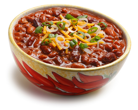 A bowl of chili beans in a bowl decorated with red chili peppers isolated on a white background.   The chili beans are topped with green onions shredded cheese.  Camera is tilted slightly off axis. A clipping path is included.