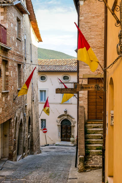 Ancient flags adorn a beautiful stone alley in a medieval town in Umbria in central Italy Ancient medieval flags adorn a beautiful stone alley in Gualdo Tadino, a medieval town in the Umbria region, in central Italy. An important city since Roman times, Gualdo Tadino rises along the ancient consular Via Flaminia, traced by the Romans. Its history runs throughout the Middle Ages and, despite having been partially destroyed and sacked numerous times and placed under the dominion of Perugia, this ancient Umbrian center still retains its medieval charm. The Umbria region, considered the green lung of Italy for its wooded mountains, is characterized by a perfect integration between nature and the presence of man, in a context of environmental sustainability and healthy life. In addition to its immense artistic and historical heritage, Umbria is famous for its food and wine production and for the high quality of the olive oil produced in these lands. Image in high definition format. gualdo tadino stock pictures, royalty-free photos & images