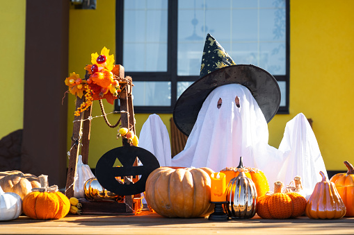 Child in bed sheet with slits like ghost in witch's hat and Halloween decor on the porch of the house outside in the yard of pumpkin, lantern, garlands, ack lantern. party, autumn mood