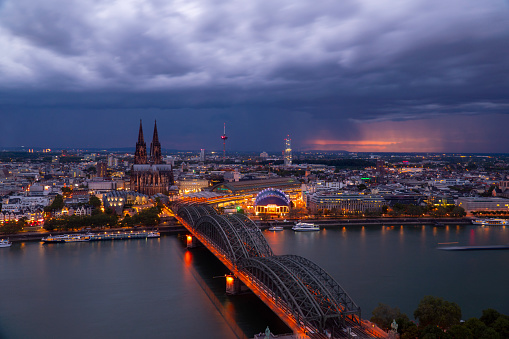 Dramatic thunderclouds over Cologne Cathedral and Hohenzollern Bridge in the evening. Artificial city lighting