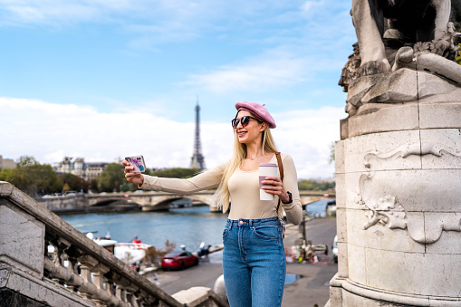 Young cheerful woman taking a selfie in front of the Eiffel Tower and River Seine in Paris, France