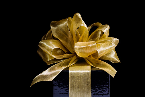 A Christmas gift wrapped in fancy, ornate, hand-tied gold ribbon and bow. Close-up of a mystery present for a special someone at a holiday event, special occasion or wedding or anniversary celebration. Isolated on black background.