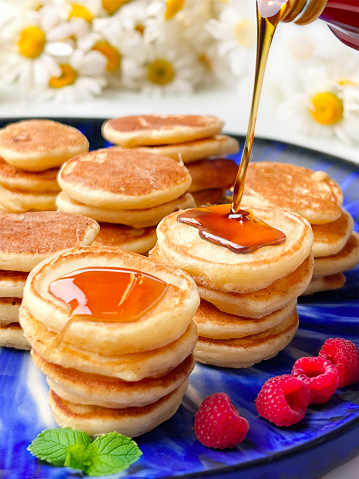 Homemade Mini Pancakes with Maple Syrup and Rasberries on a Blue Plate and Fresh Chamomilles Background.