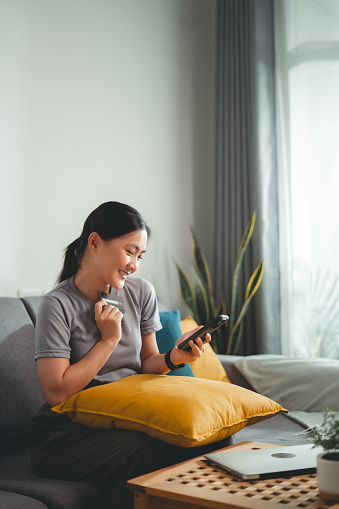 Asian woman sitting on sofa in living room using smart phone and credit card for shopping online at home. Happy excited woman shopper using instant mobile payments making purchase in online store.