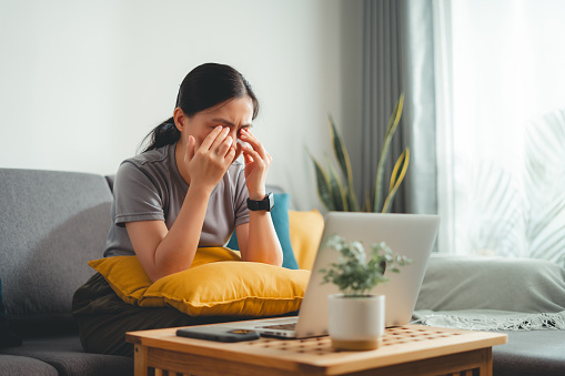 Asian woman feel eye pain while using laptop, rubbing irritated eyes sitting on sofa at home.