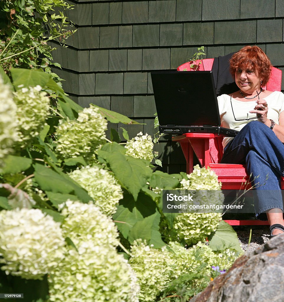 Laptop In The Garden Woman in garden with laptop. Holding Sun glasses and sitting in Red Recycled Plastic Adirondack Chair. Anabelle Hydrangeas in foreground. Green shingle siding in background. Siding - Building Feature Stock Photo