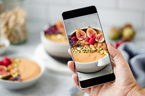 Point of view of a woman photographing a healthy smoothie bowl on kitchen counter with her mobile phone. Close-up of female hand holding smartphone and taking photos freshly make breakfast smoothie on table.
