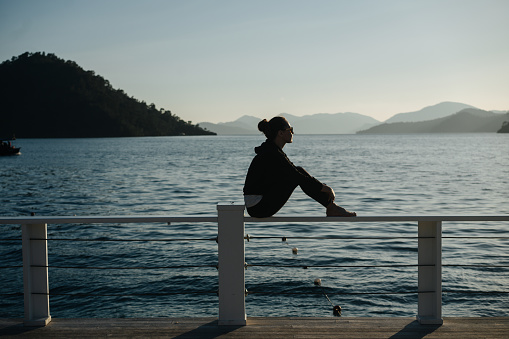 Silhouette of a girl sitting on the railing hugging her knees against the backdrop of the sea