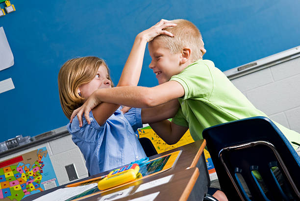 A pair of primary school boys fight playfully in the classroom while the teacher turns to take control of the situation.Candid, horizontal group portrait.