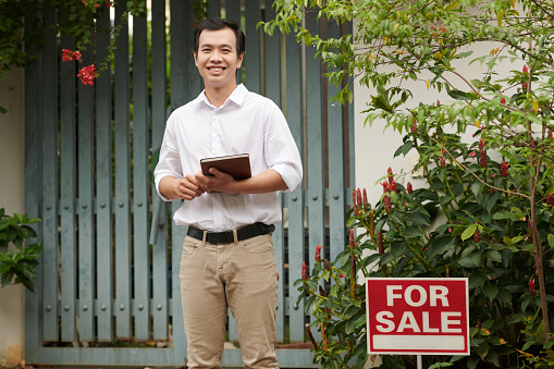 Portrait of smiling real estate agent with tablet computer standing at gate of house on sale