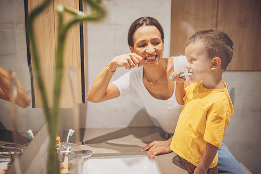Little boy brushing teeth in domestic bathroom, his mother is teaching him.