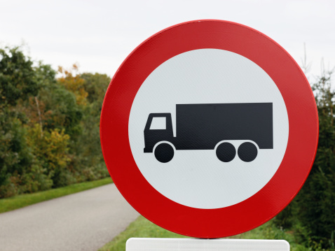 Close-up of a round red & white road sign depicting a black truck alongside a road. Focus is on the sign.