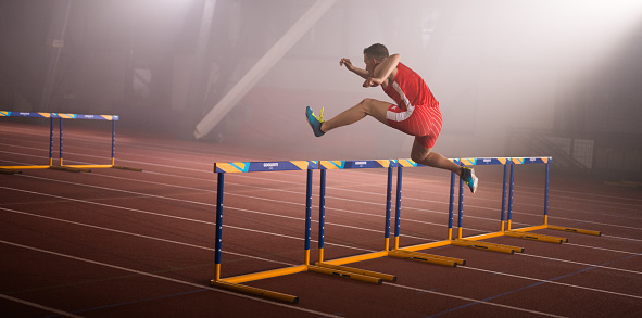 Young male runner training and jumping over hurdles at indoors stadium running track. Sport and competition concept.