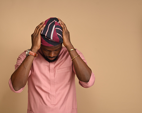 Portrait of Happy Handsome black Man holding his head as he adjusts his African cap in Cozy studio at Home Standing on brown backdrop. Close-up Profile Shot.